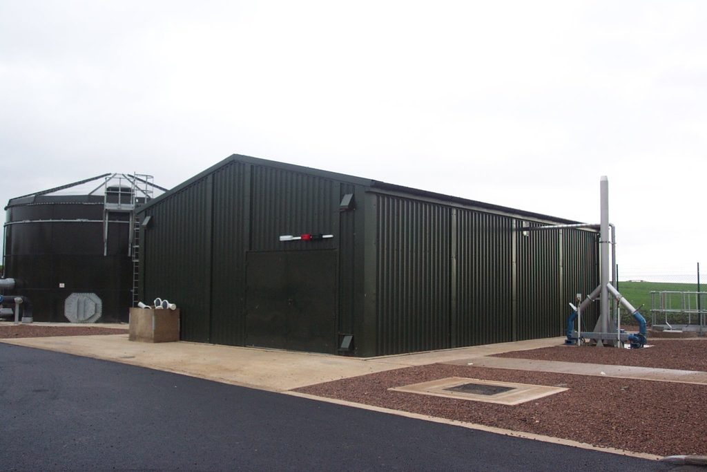 Dark green prefabricated steel wastewater treatment works building with large access door and external pipework.