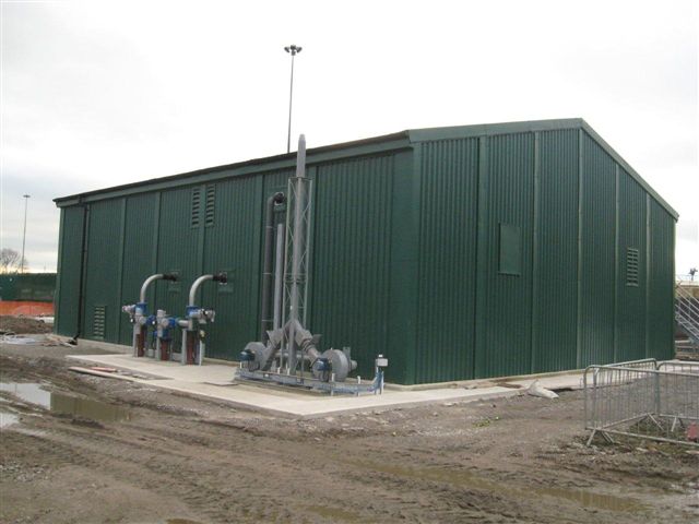 Prefabricated steel wastewater treatment works building with dark green cladding, external pipework and pumping equipment.
