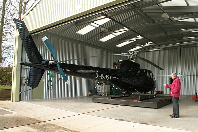 Helicopter housed inside a steel aircraft hangar with rooflights and wide access opening.